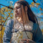 A woman with tattoos poses in a vintage dress surrounded by natural branches outdoors.
